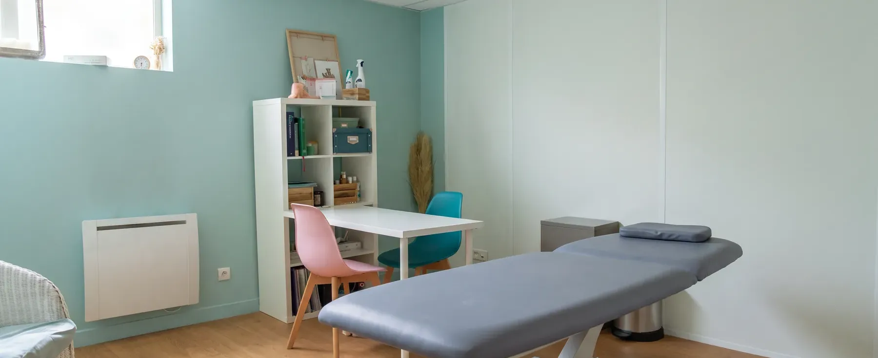 La salle de soin en lumière du jour, avec son mur vert d’eau, son étagère blanche, ses chaises rose et bleu canard et la table de soin grise au premier plan.