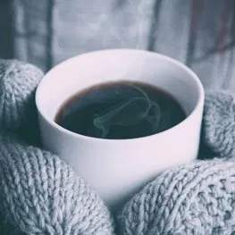 Une tasse de café fumant, tenue entre deux mains en moufles de laine.