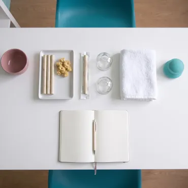 Vu de dessus, sur le bureau blanc du cabinet : une coupelle blanche avec trois bâtons de moxa et un peu d’armoise séchée, un bâton de moxa sous emballage, deux ventouses en verre, une serviette pliée et un carnet ouvert avec un stylo.
