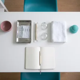 Vu de dessus, sur le bureau blanc : un carnet ouvert avec un stylo, un plateau d’aiguilles sous blister, un bâton de moxa et deux ventouses.