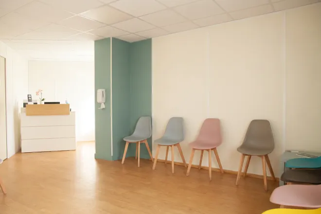 La salle d’attente du cabinet en lumière naturelle, avec sa rangée de chaises grises, rose et taupe, son poteau vert sauge et l’accueil en bois au fond.